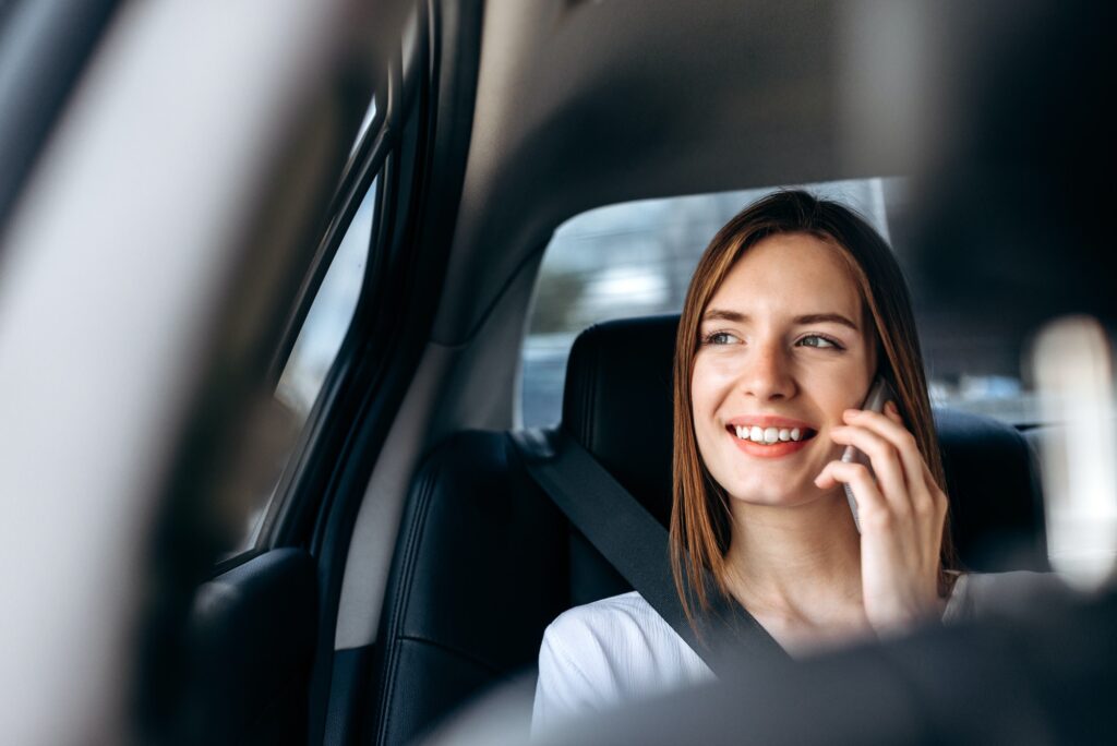 Young smiling woman holding smartphone in her hand and talking on the back seat in car